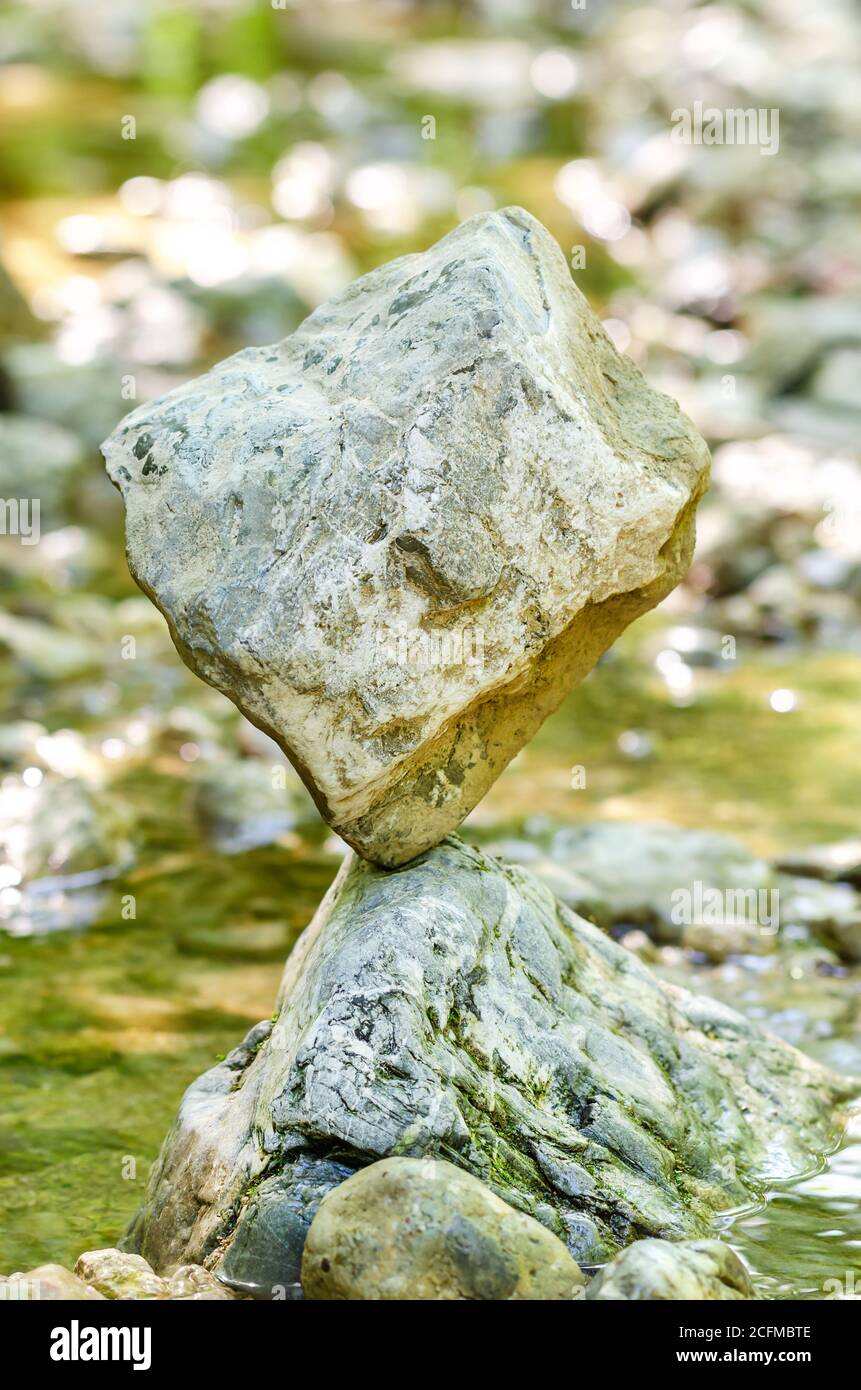 Ausgewogener Felsen am Bach. Ein würfelförmiges Gestein wird natürlich auf einem Felsen in einem Bach, der von Wasser umgeben ist, ausgeglichen. Klassische Steinbalance. Stockfoto