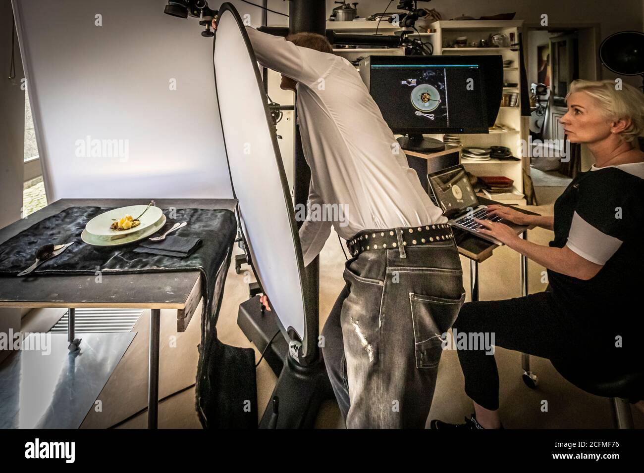 Bui Vössing lehnt sich in einen weißen Lichtdiffusor über dem Fototisch, während Su Vössing das Ergebnis des Aufnahmes auf dem Laptop überprüft, im Fotostudio in Stolberg, Deutschland. Stockfoto