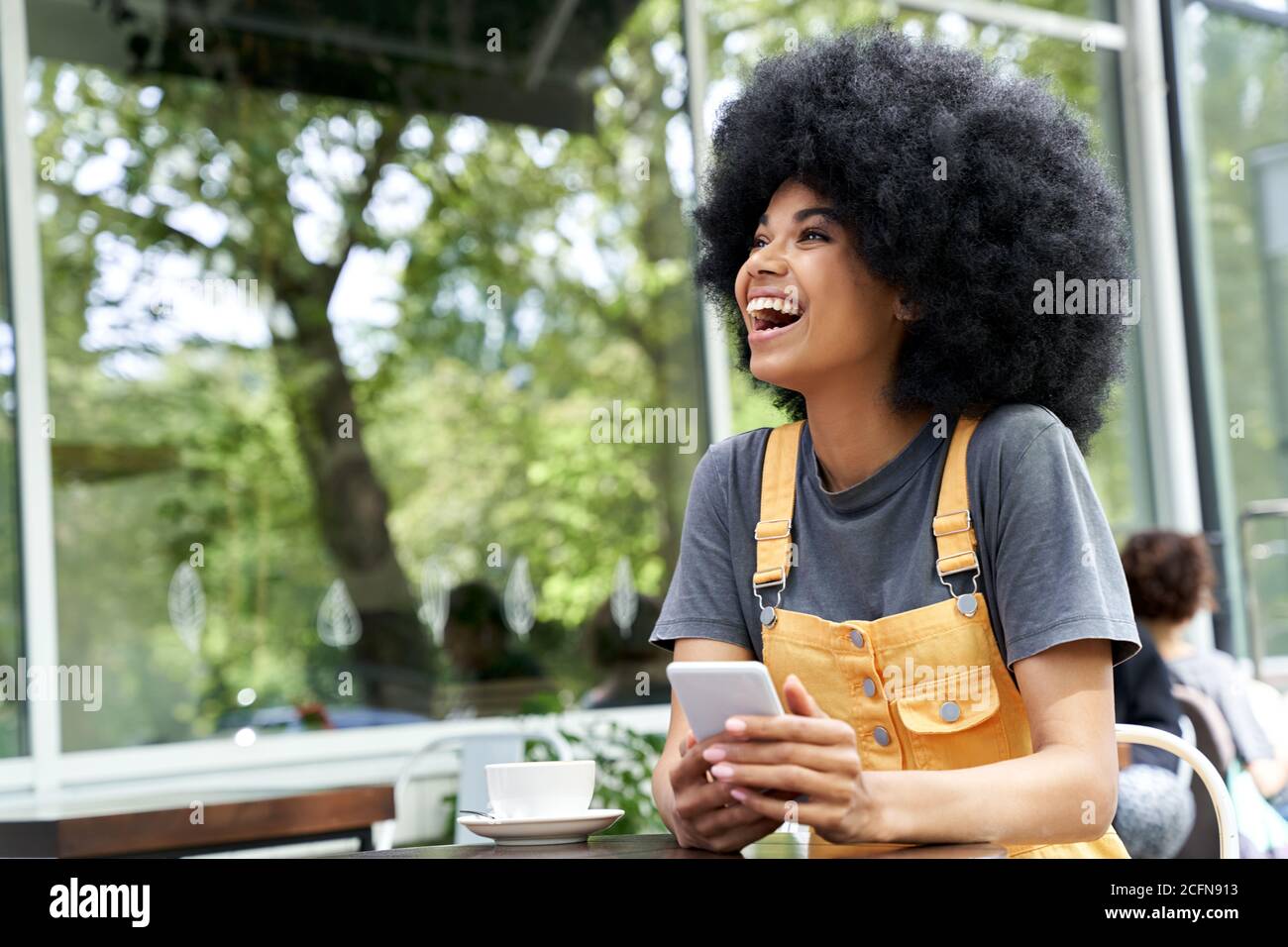 Fröhliche afrikanische Frau, die Telefon benutzt, lachend, am Kaffeetisch im Freien sitzend. Stockfoto