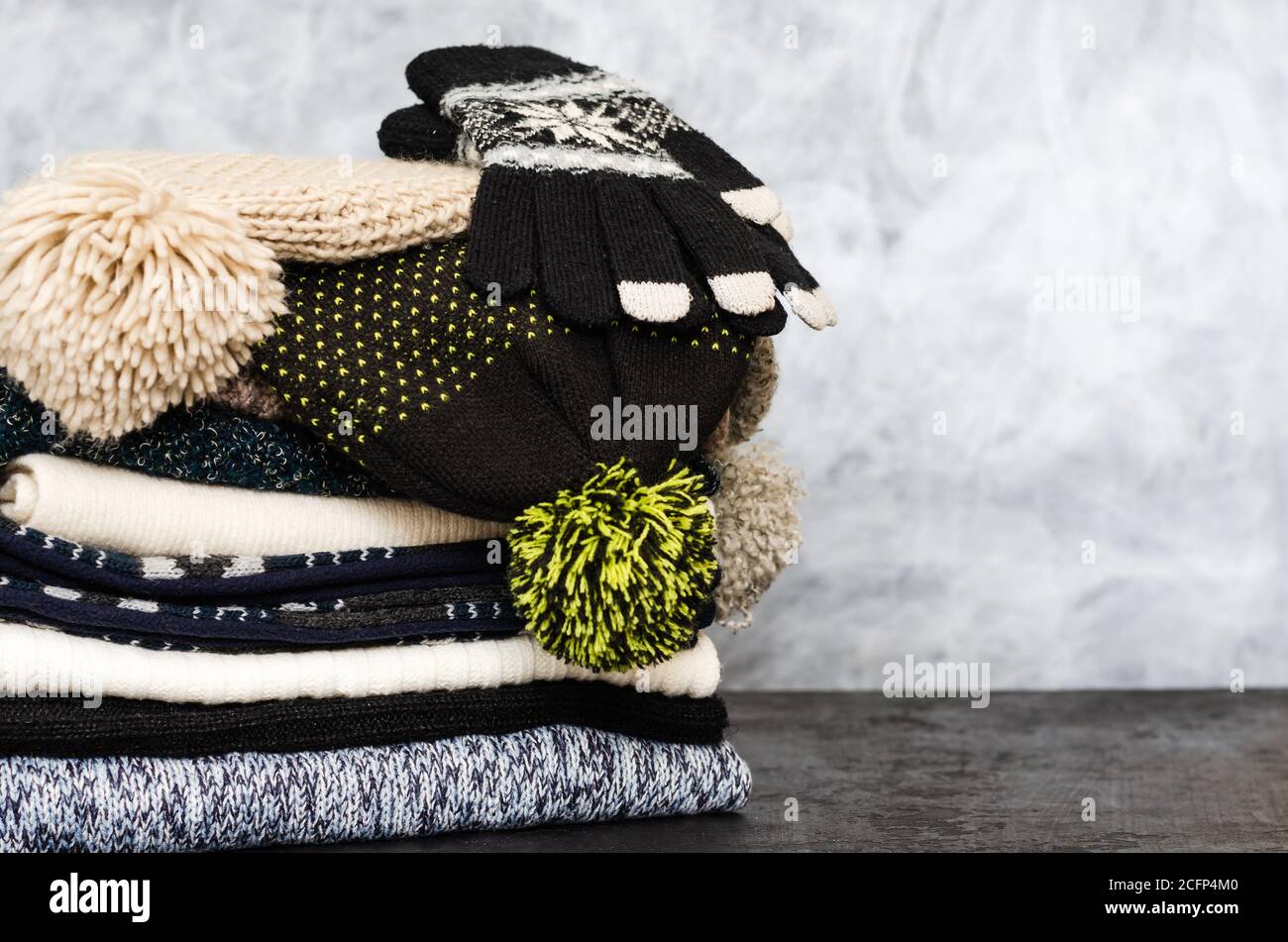 Ein Stapel von ordentlich gefalteten gestrickten Winterschals, Hüten mit Pompons und Handschuhen auf grauem Hintergrund. Saison der warmen Kleidung. Zeit zum Warmhalten. Nahaufnahme Stockfoto