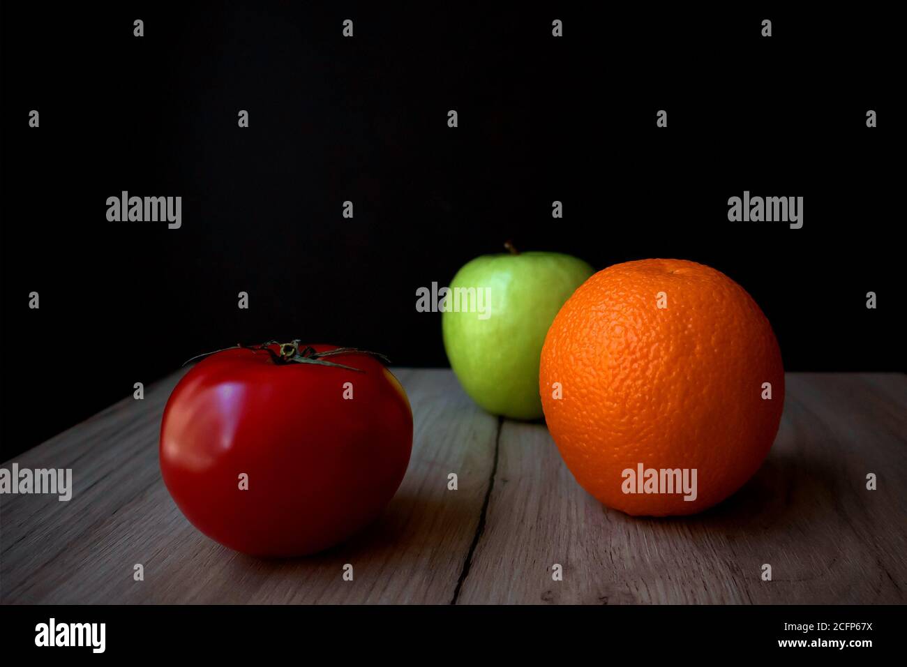 Komposition aus reifen Tomaten, grünem Apfel und Orange, die auf einem Holzbrett vor schwarzem Hintergrund liegen Stockfoto