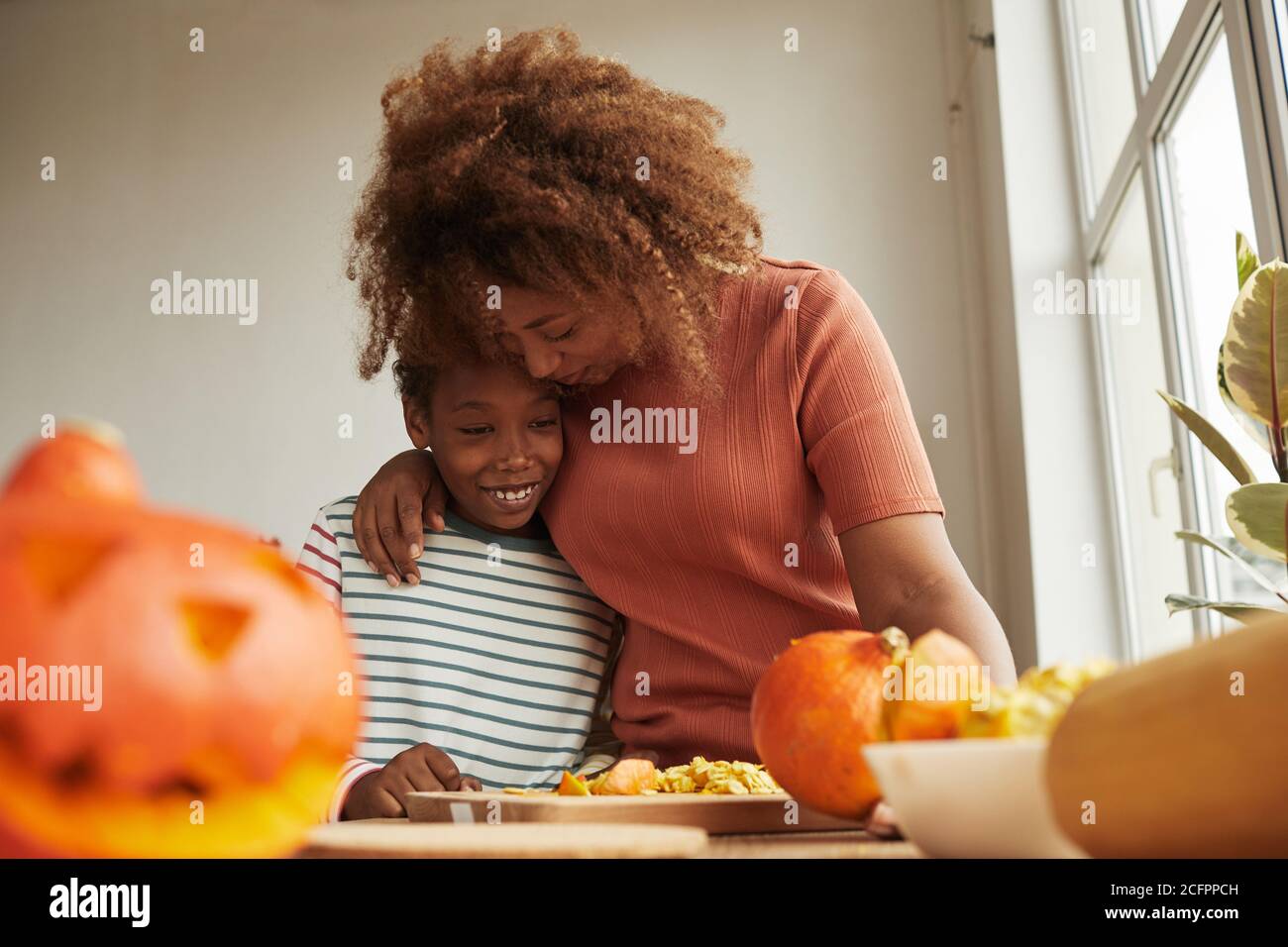 Liebevolle Mutter umarmt ihren fröhlichen Sohn, während sie reife Kürbisse schnitzt Für Halloween Party zusammen Stockfoto