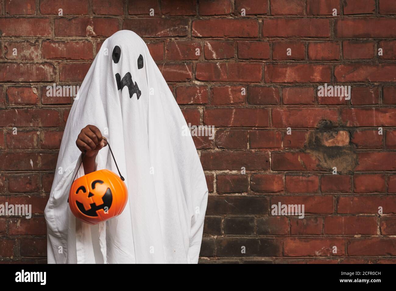 Nicht erkennbares Kind verkleidet als Geist trägt weißen Stoff halten jack o' Laterne Korb für Süßigkeiten steht gegen Backsteinmauer Stockfoto