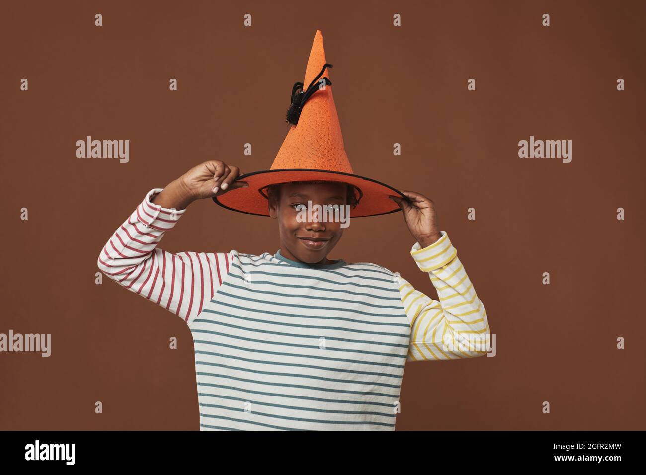 Studioportrait eines fröhlichen Jungen, der an einer braunen Wand steht Hintergrund versucht auf lustige orange Zauberer Hut Blick auf Kamera Stockfoto