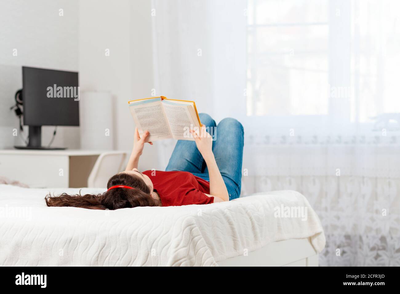 Eine junge kaukasische Frau liegt auf dem Bett auf ihrem Rücken und liest ein Buch. Speicherplatz kopieren. Weißer Innenraum des Raumes auf dem Hintergrund. Konzept der Bildung Stockfoto