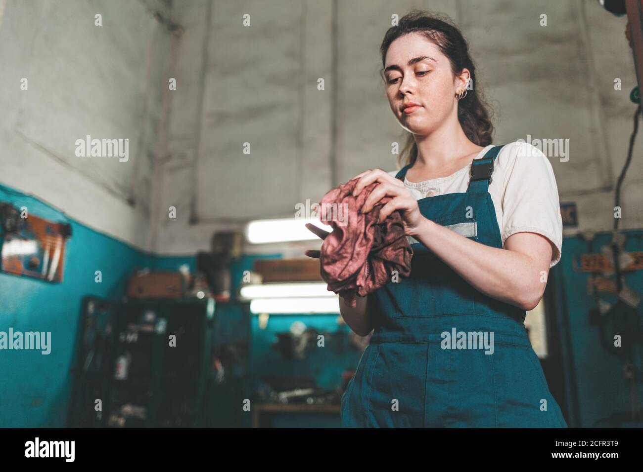 Gleichstellung der Geschlechter. Portrait einer jungen Frau in Uniform, die in einer Werkstatt arbeitet, die seine Hände mit einem Lappen abwischt. Ansicht von unten. Autogeschäft im Hintergrund Stockfoto
