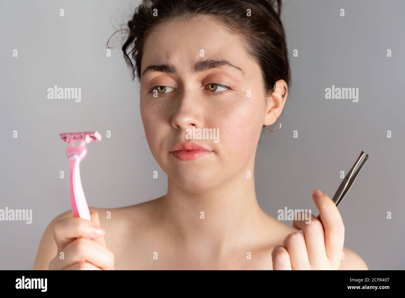 Eine junge hübsche Frau hält ein Paar Pinzette und ein Rasiermesser, nicht zu wissen, wie überschüssige Haare auf ihrem Gesicht zu entfernen.das Konzept der loszuwerden unerwünschte Stockfoto