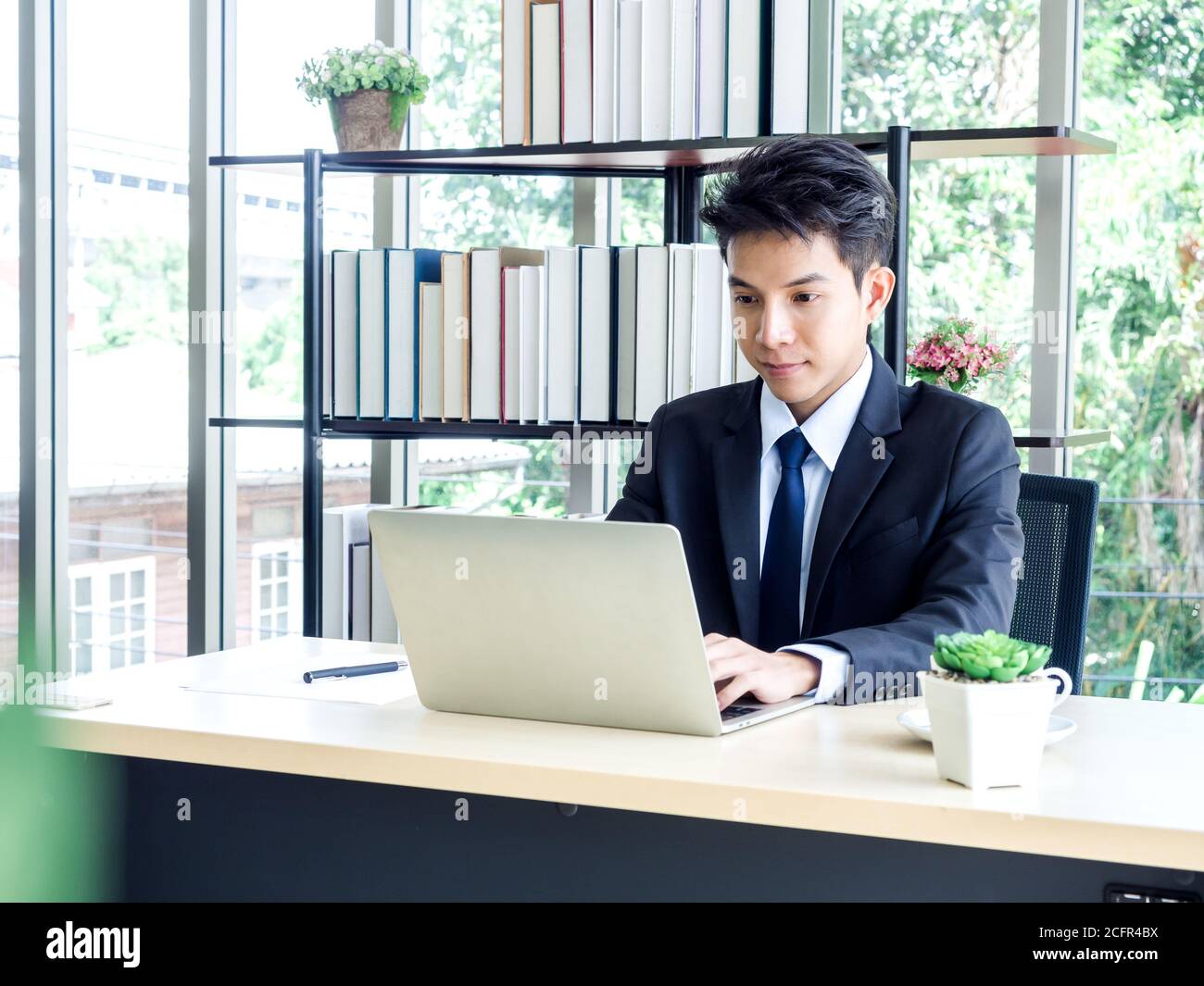 Junger asiatischer Geschäftsmann im Anzug mit Laptop-Computer im Büro mit grünem Baum Hintergrund draußen arbeiten. Mann im Anzug mit Laptop in gut beleuchteten Arbeitsplatz Stockfoto