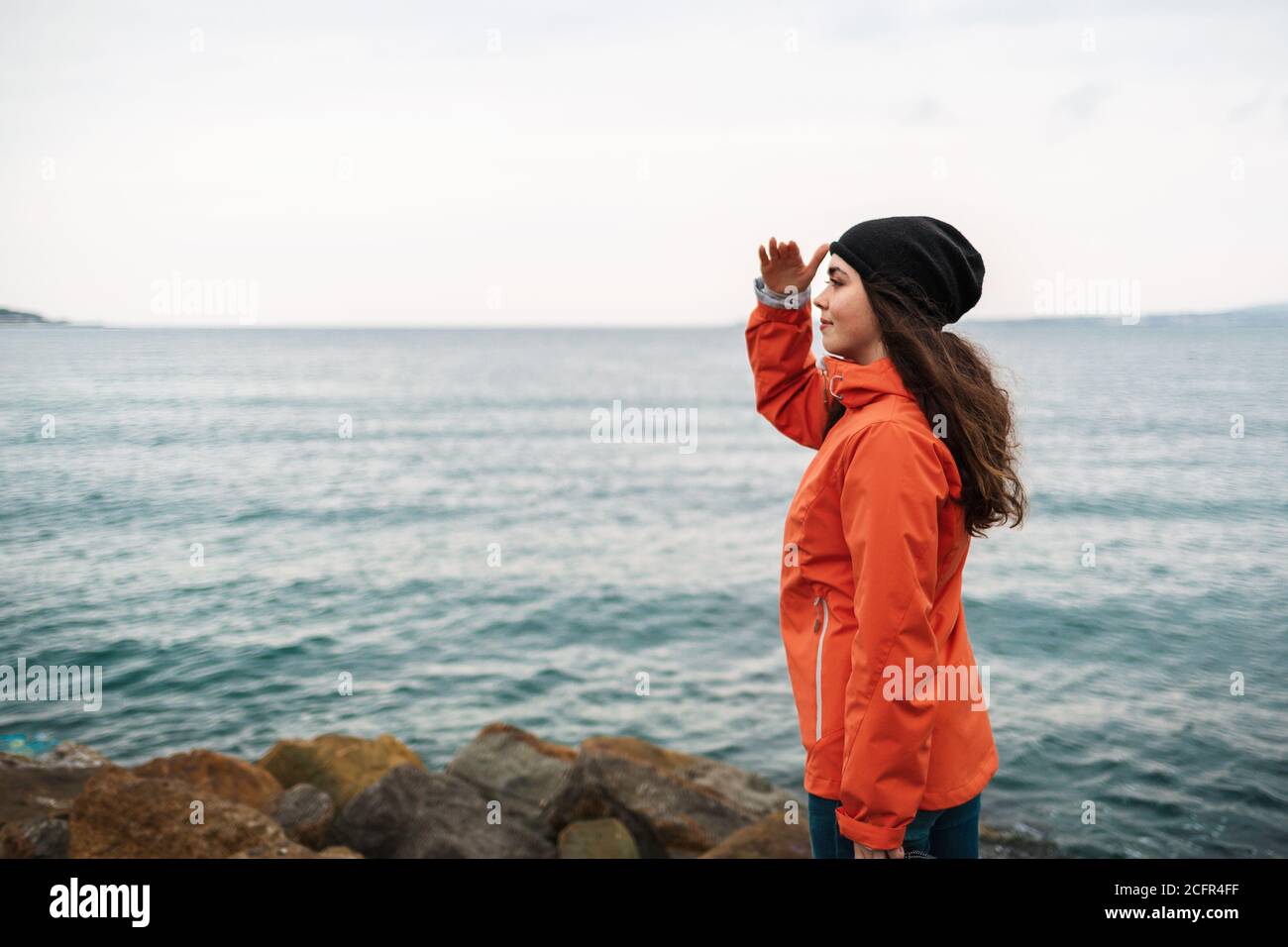 Eine Brünette mit langen Haaren, die eine Mütze und eine orangefarbene Jacke trägt, steht am Ufer und schaut in die Ferne. Im Hintergrund das Meer und die Stockfoto