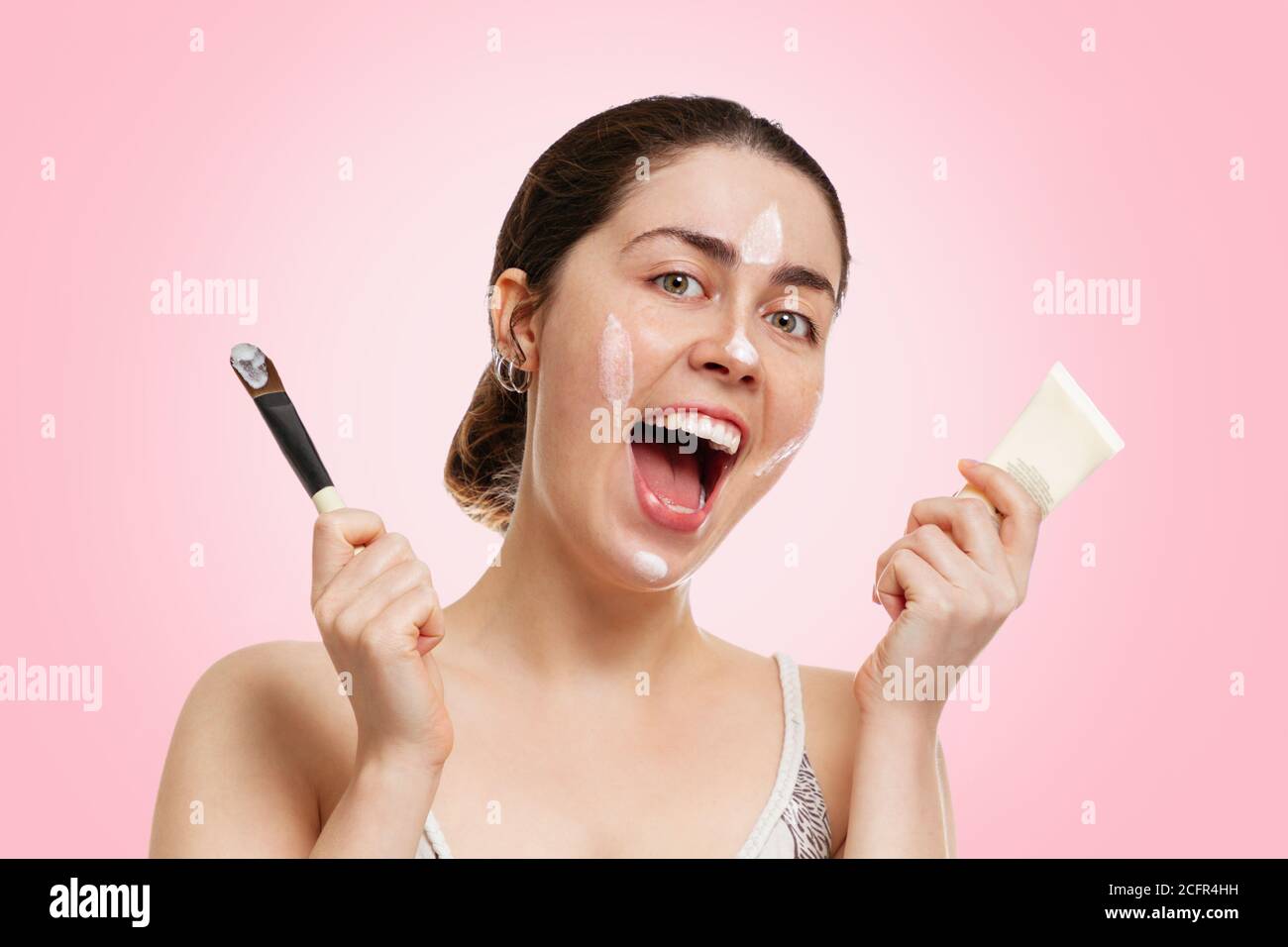 Porträt einer jungen schönen Frau mit einem glücklichen Lächeln, hält einen Schlauch mit einer Maske und einem Pinsel in der Hand. Rosa Hintergrund. Konzept der Selbstversorgung, cos Stockfoto