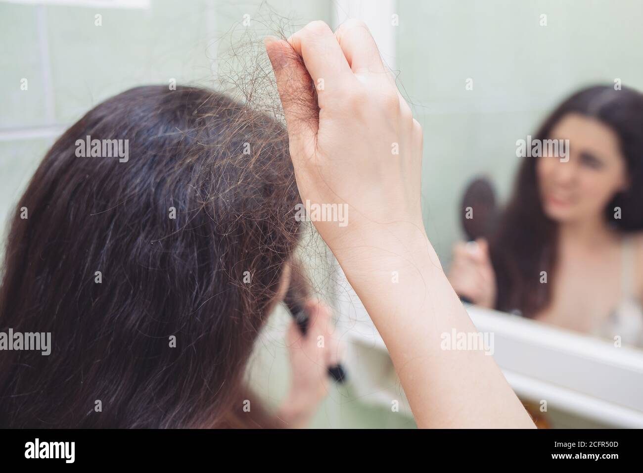 Eine Brünette zieht ein paar Haare aus ihrem Kopf. Rückansicht. Im Hintergrund eine Spiegelung im Spiegel. Das Konzept des Haarausfalls. Stockfoto