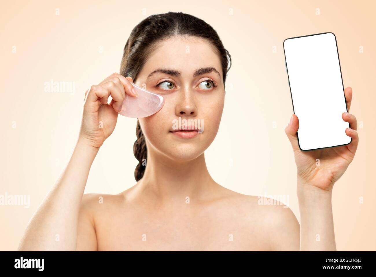 Gua Sha Massage. Eine junge Frau hält ein Handy in der einen Hand und einen Quarzstein Gua Sha in der anderen. Modell. Beigefarbener Hintergrund. Geschäftskonzept Stockfoto