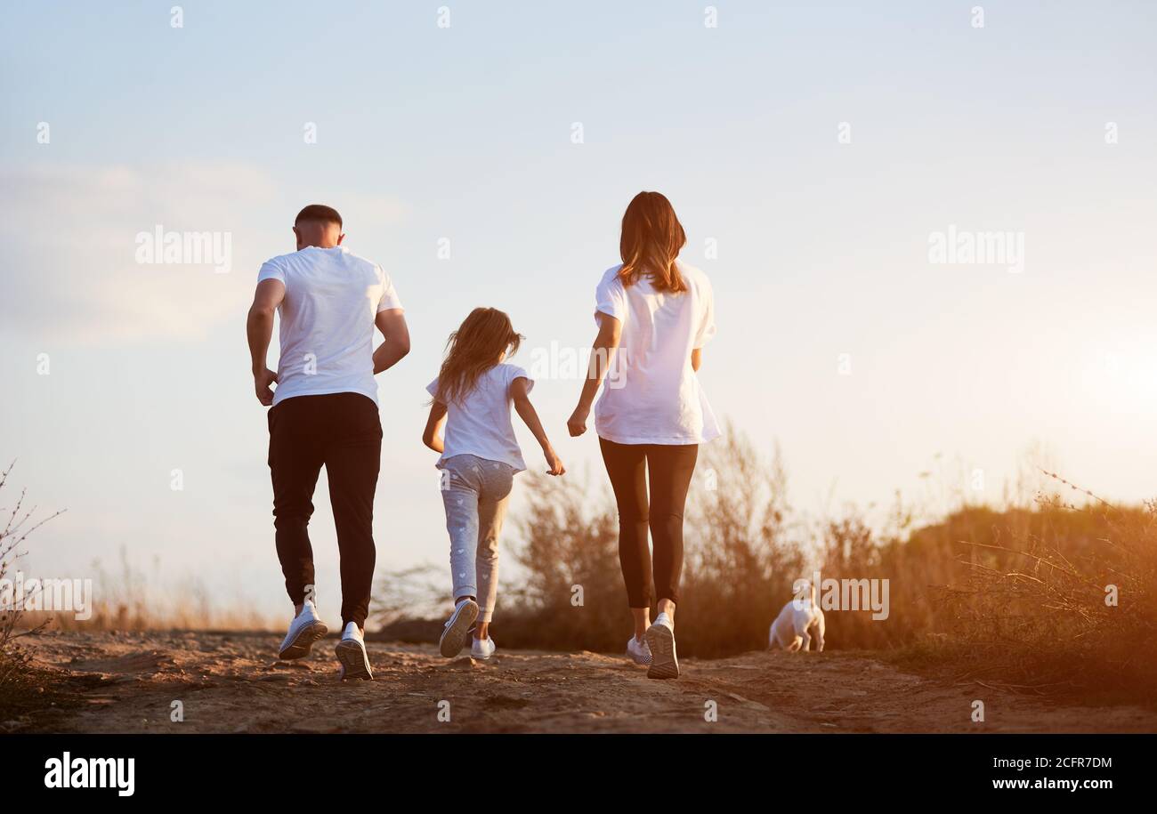 Rückansicht der sportlichen und schönen Familie von drei Joggen Mit ihrem Jack russell Terrier außerhalb der Stadt auf der Dorfstraße bei Sonnenuntergang Stockfoto