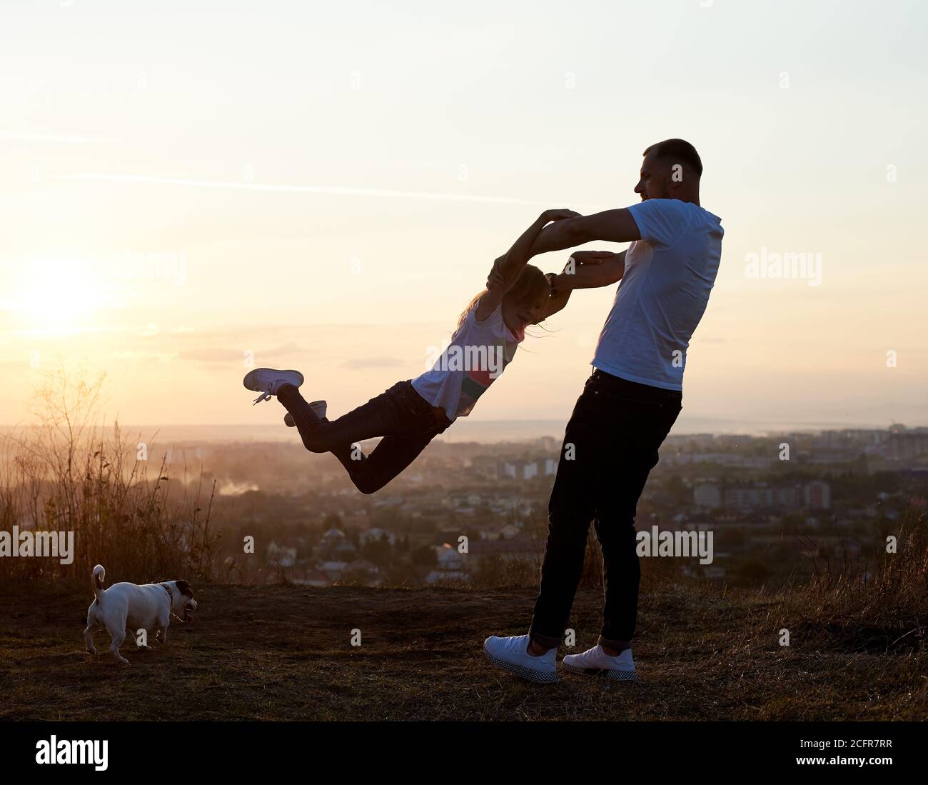 Silhouette eines Vaters schwingt seine Tochter an den Armen auf dem Hügel auf der untergehenden Sonne in den Vororten, Jack russell Terrier in ihrer Nähe, schöner Horizont im Hintergrund, Seitenansicht Stockfoto
