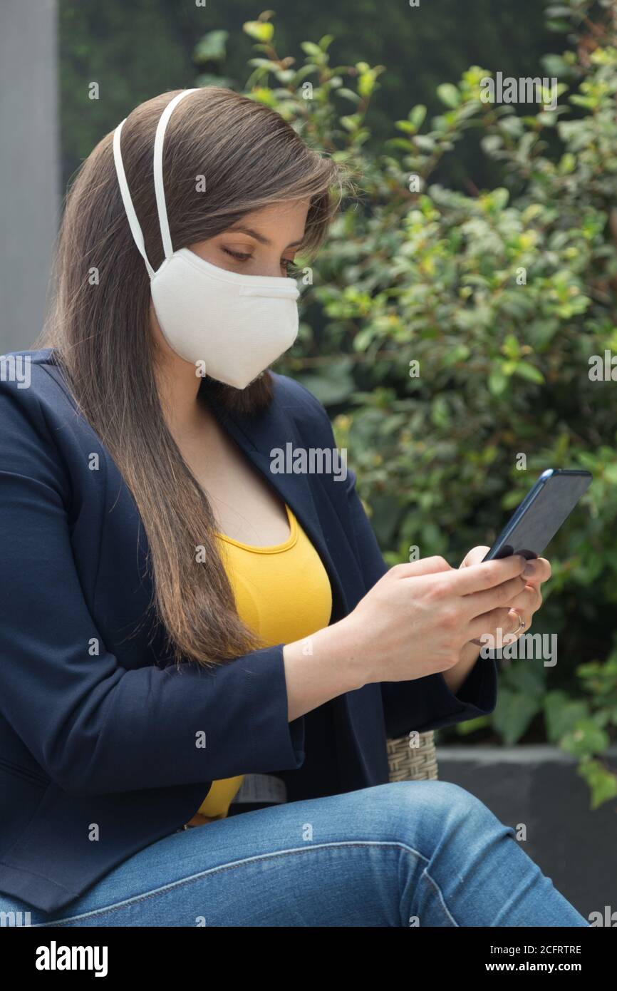 Junge Frau mit langen Haaren und beobachten ihr Handy, trägt eine gelbe Bluse, blaue Jacke und Maske Stockfoto