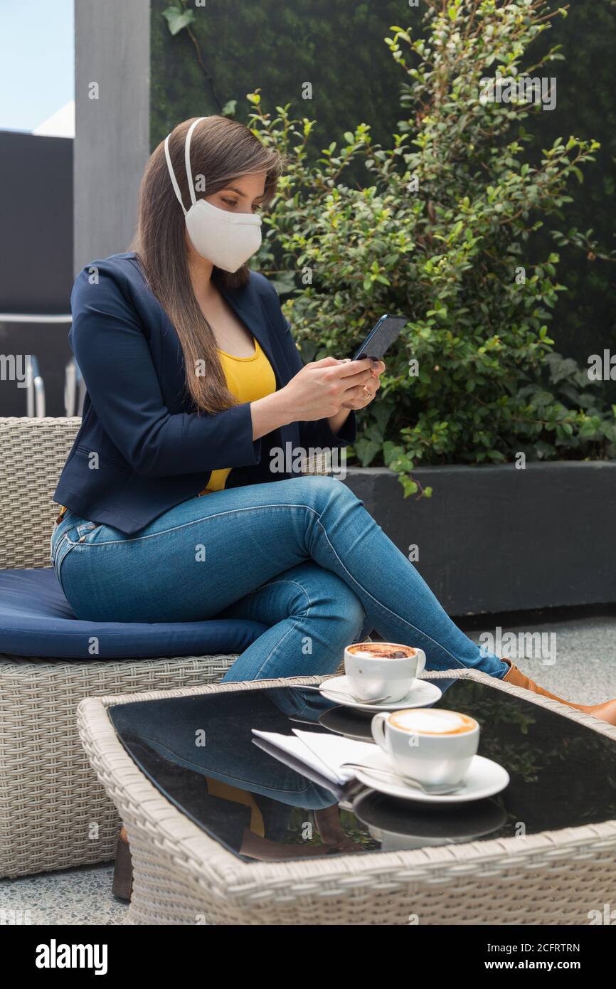 Junge Frau mit langen Haaren sitzt draußen neben einigen Tassen Kaffee, trinken und auf ihr Handy, in einer gelben Bluse, blaue Jacke A Stockfoto