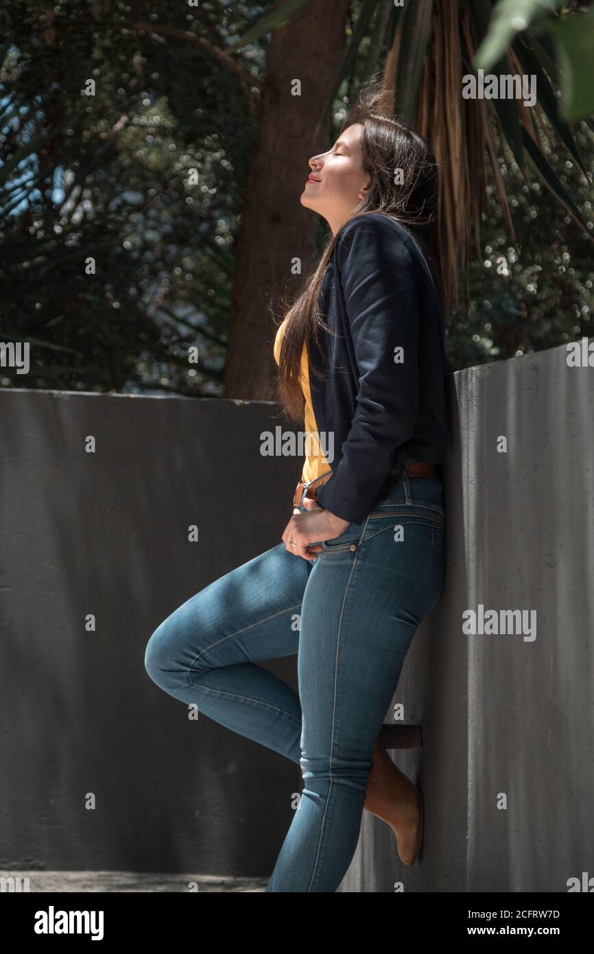 Junge Frau im Profil mit langen Haaren im Schatten stehend, lächelnd und posiert, verwendet Jeans und schwarze Jacke Stockfoto