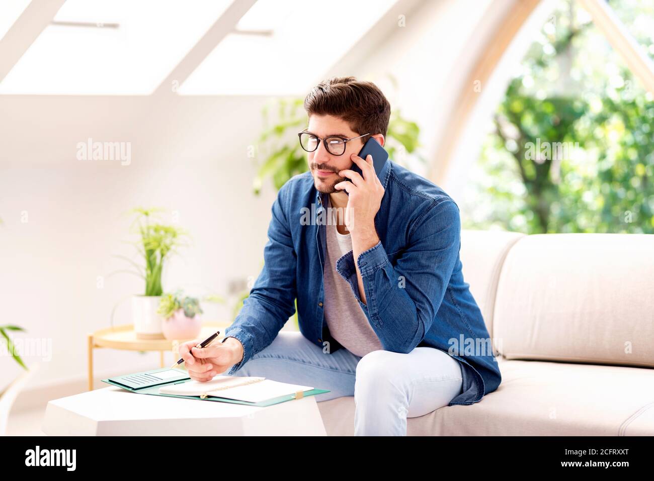 Aufnahme eines Geschäftsmannes, der einen Anruf macht und in seinen Ordner schreibt, während er zu Hause auf dem Sofa sitzt. Stockfoto