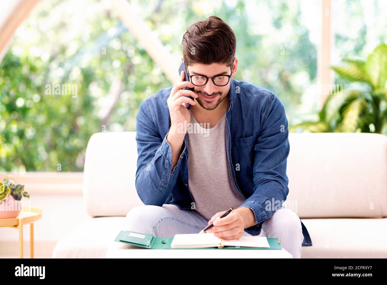 Aufnahme eines Geschäftsmannes, der einen Anruf macht und in seinen Ordner schreibt, während er zu Hause auf dem Sofa sitzt. Stockfoto