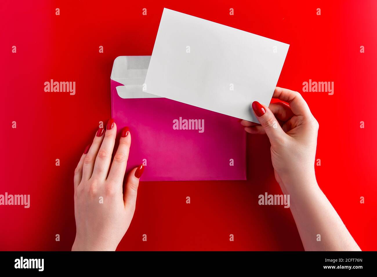 Ein Gratulationsumschlag in den Händen einer jungen Frau auf rotem Hintergrund. Layout für Feiertagsgrüße. Rosa Umschlag auf rotem Hintergrund, Platz fo Stockfoto