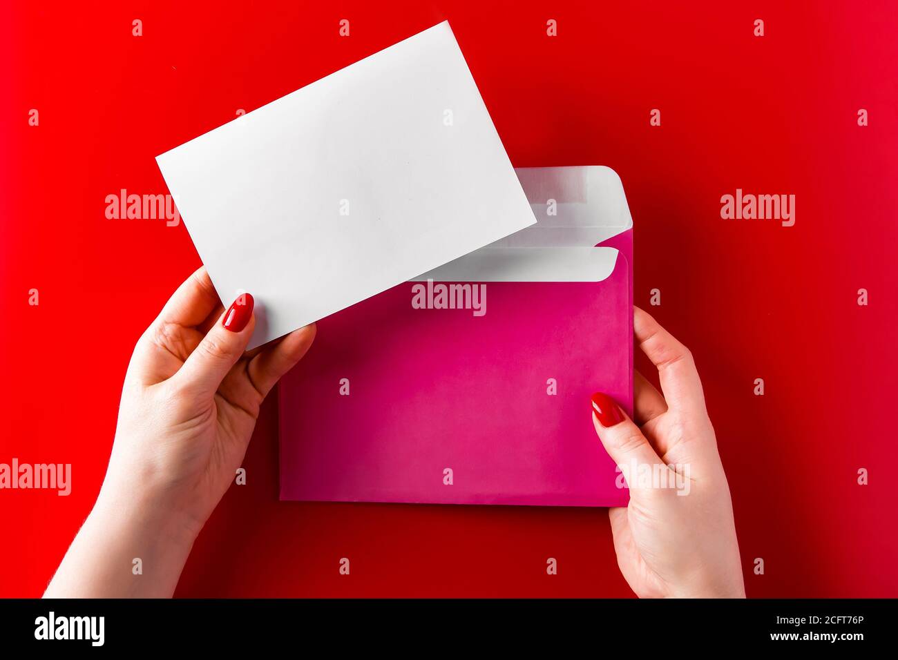 Ein Gratulationsumschlag in den Händen einer jungen Frau auf rotem Hintergrund. Pinkfarbener Umschlag und Grußkarte in Händen auf rotem Hintergrund. Stockfoto