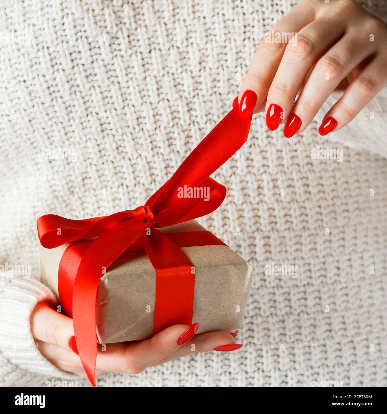 Geschenk mit einem roten Band in den Händen auf weißem Hintergrund. Eine Frau packt eine Schachtel mit einer Überraschung aus. Ein Geschenk für den Urlaub. Stockfoto