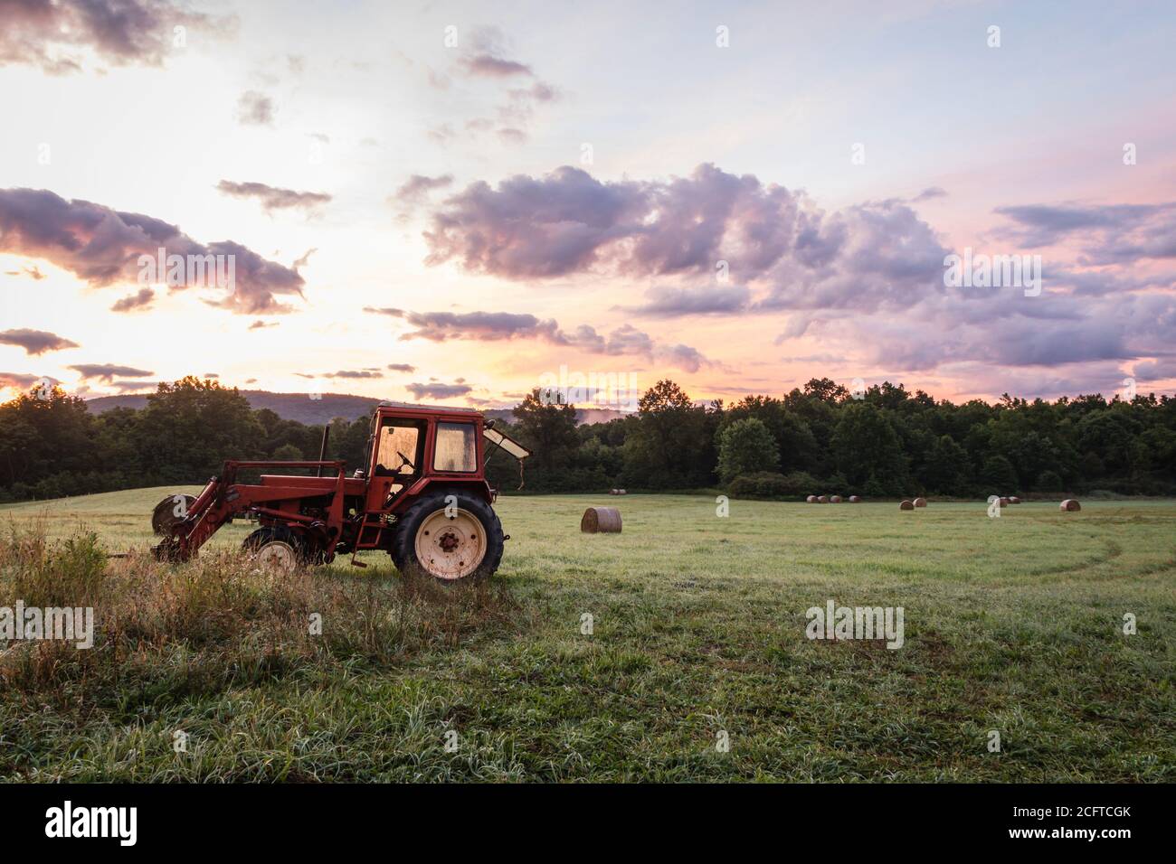 Roter Traktor und frisch gerollte Heuballen ruhen auf Rollen Hügel mit dramatischer Wolkenlandschaft bei Sonnenaufgang schaffen eine rustikale ländliche Szene Stockfoto