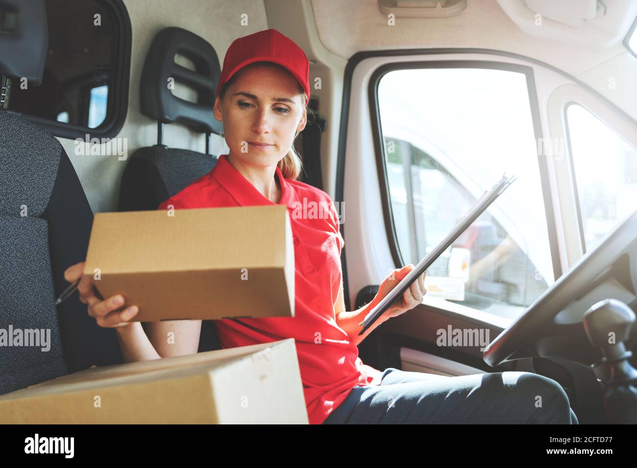 Weibliche Lieferdienstarbeiterin in roter Uniform sitzt im Lieferwagen Und mit Paketen arbeiten Stockfoto