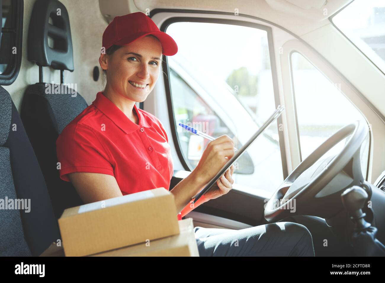Junge lächelnde Delivery Person in roter Uniform sitzt in van Und das Schreiben von Dokumenten Stockfoto