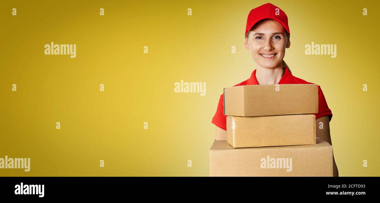 Lieferservice - junge lächelnde deliverywoman in roten Uniform halten Kartons vorne auf gelbem Hintergrund mit Kopierplatz Stockfoto