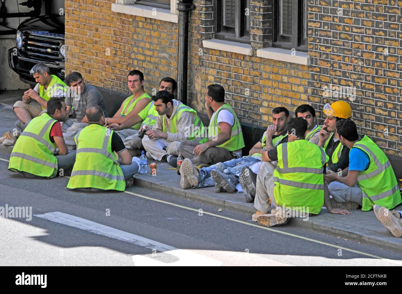 Londoner Straßenszene Gruppe von Baustellenarbeitern im Schatten auf dem Bürgersteig außerhalb der Baustelle in Warnjacken zur Mittagszeit in Großbritannien Stockfoto