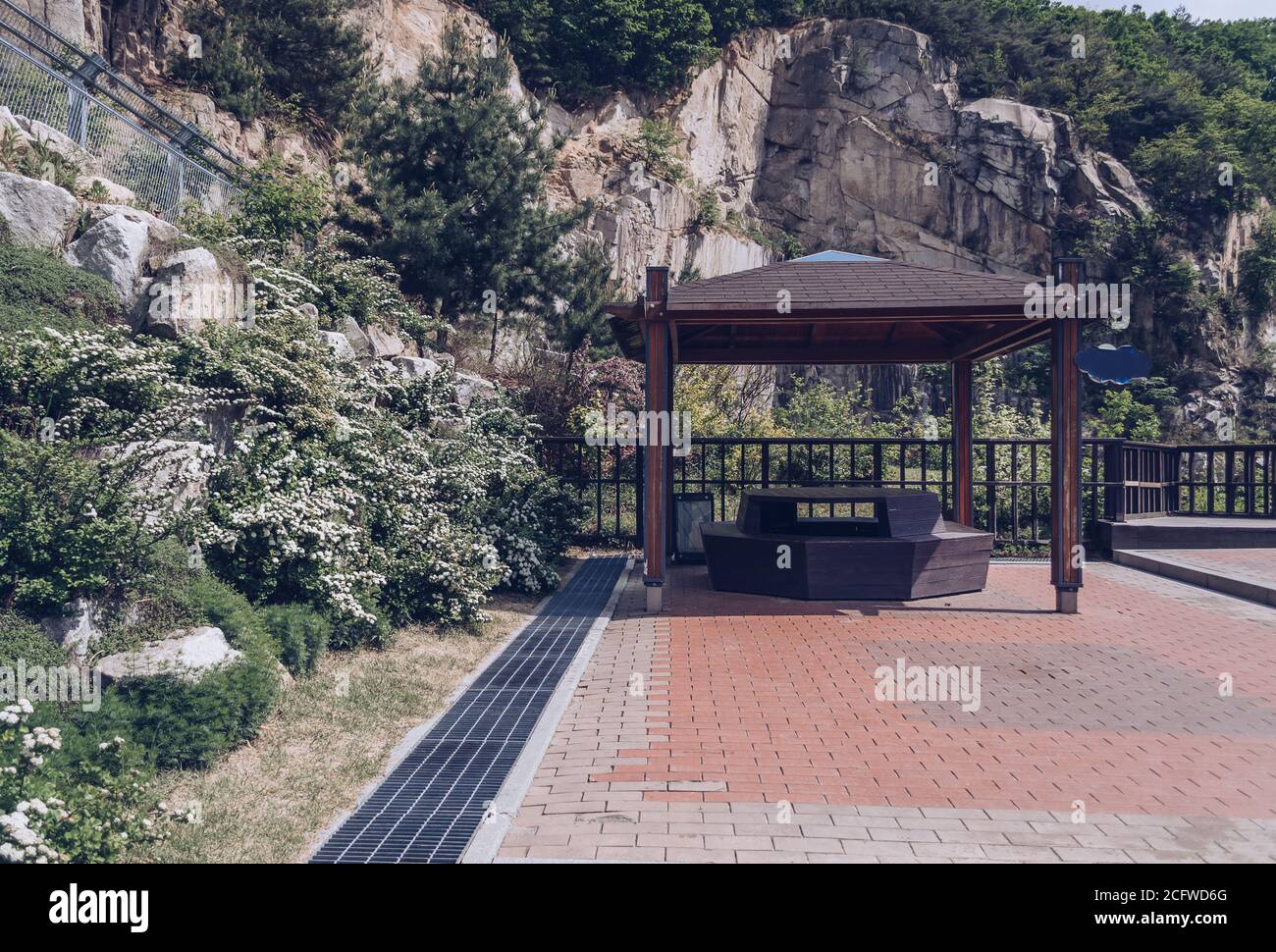 koreanischer Pavillon aus Holz mit Büschen mit Spiraea-Blüten an der Seite Und Klippe bedeckt mit Bäumen auf der anderen Stockfoto