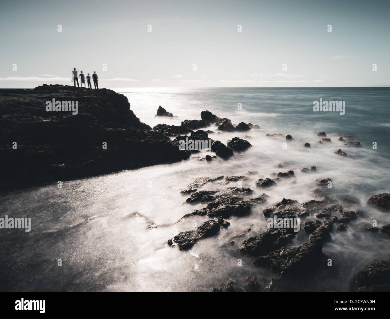 Eine lange Exposition von vier Menschen auf einer Klippe stehen Am Meer mit Felsen und Wellen im Vordergrund Und der Horizont und der Himmel im Hintergrund Stockfoto