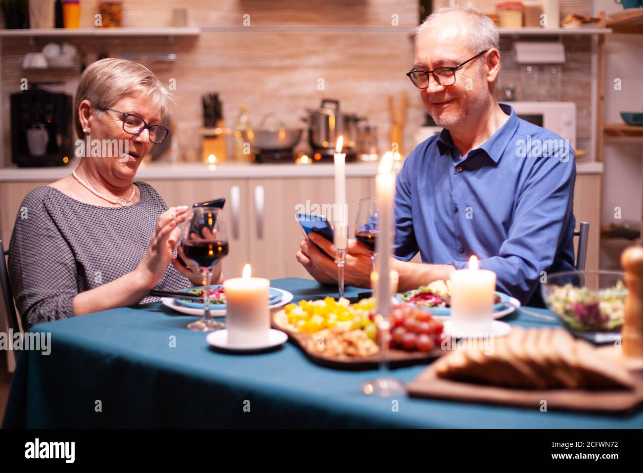 Glückliches altes Paar, das lächelte und in der Küche ein Telefon benutzte, um ein romantisches Abendessen zu genießen. Sitzen am Tisch im Speisesaal, Surfen, Suchen, Telefon, Internet, ihren Jahrestag zu feiern Stockfoto