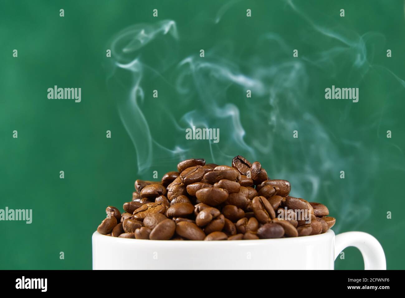Internationaler Tag des Kaffees Konzept. Nahaufnahme weiße Kaffeetasse voller Kaffeebohnen auf grünem Hintergrund mit Rauch auf der Oberseite. Stockfoto