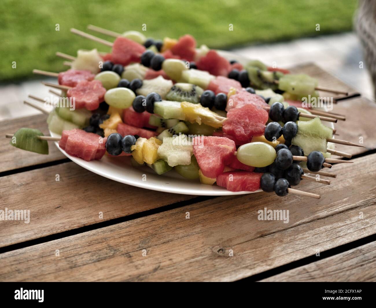 Frisches Obst auf Holzstäbchen bereit Tisch für Gartenparty. Heidelbeeren, Trauben, Kiwi, Ananas, Wassermelone. Verschwommenes grünes Gras auf dem Hintergrund. Stockfoto