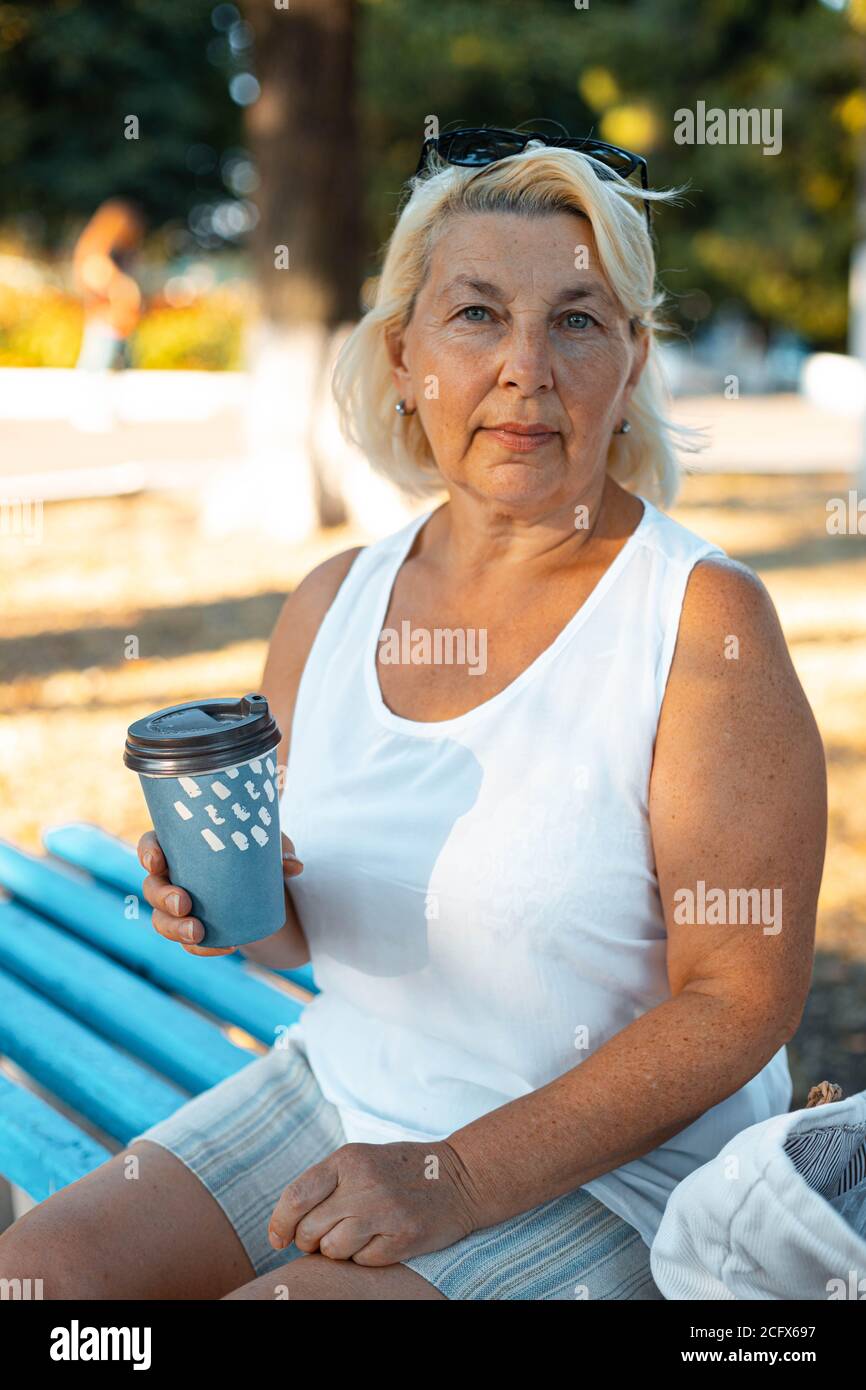 Erwachsene blonde lächelnde Frau mit Tasse Kaffee zum Mitnehmen sitzt auf ood Bank im grünen Park am Sommertag, Stockfoto