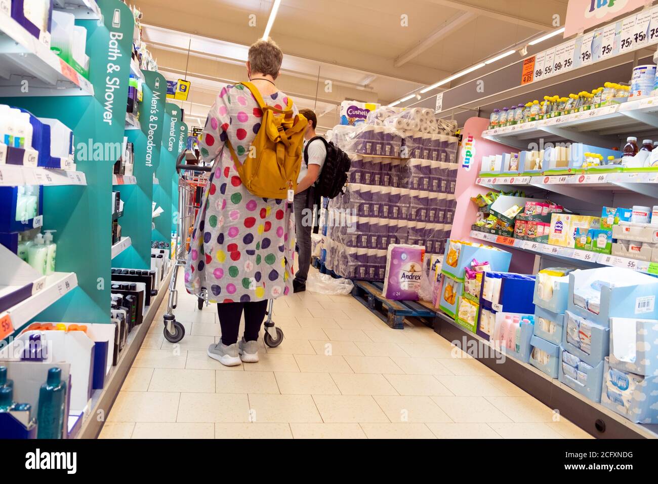 Stilvolle Frau trägt bunte Polka Dot Mantel & gelben Rucksack Rucksackeinkauf in Lidl Supermarkt stehende Papierprodukte Gang UK KATHY DEWITT Stockfoto