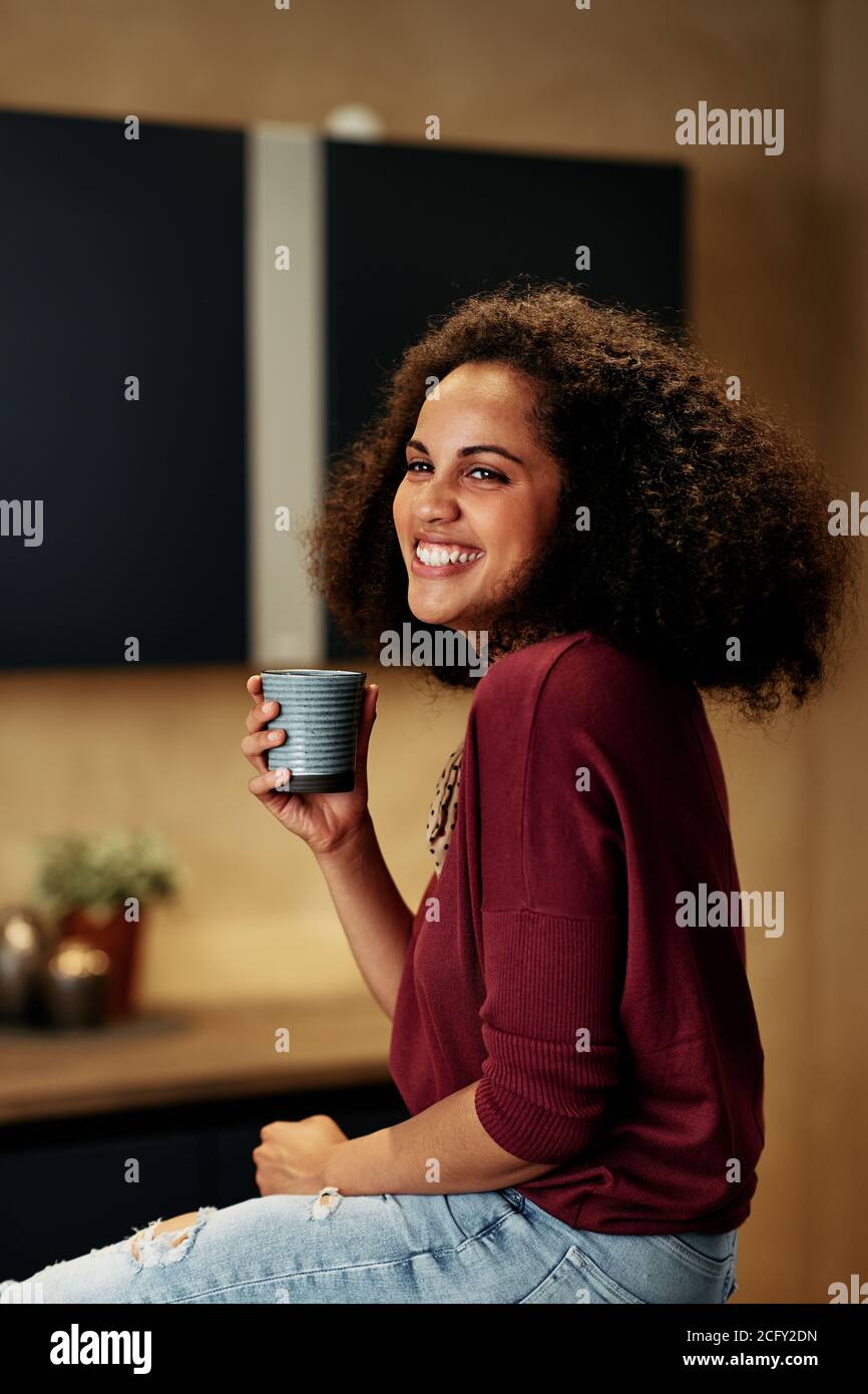 Lächelnde junge afroamerikanische Frau, die auf einer Theke sitzt Ihre Küche und eine Tasse Kaffee trinken Stockfoto