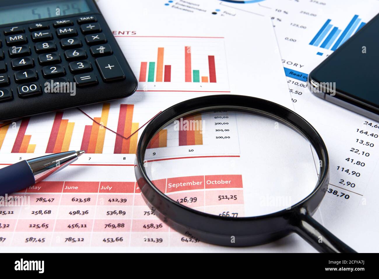 Ansicht des Büroarbeitsplatzes mit Finanzkarten und Zahlen, mit Taschenrechner, Lupe und Mobiltelefon. Blauer Stift auf weißem Papier. Stockfoto