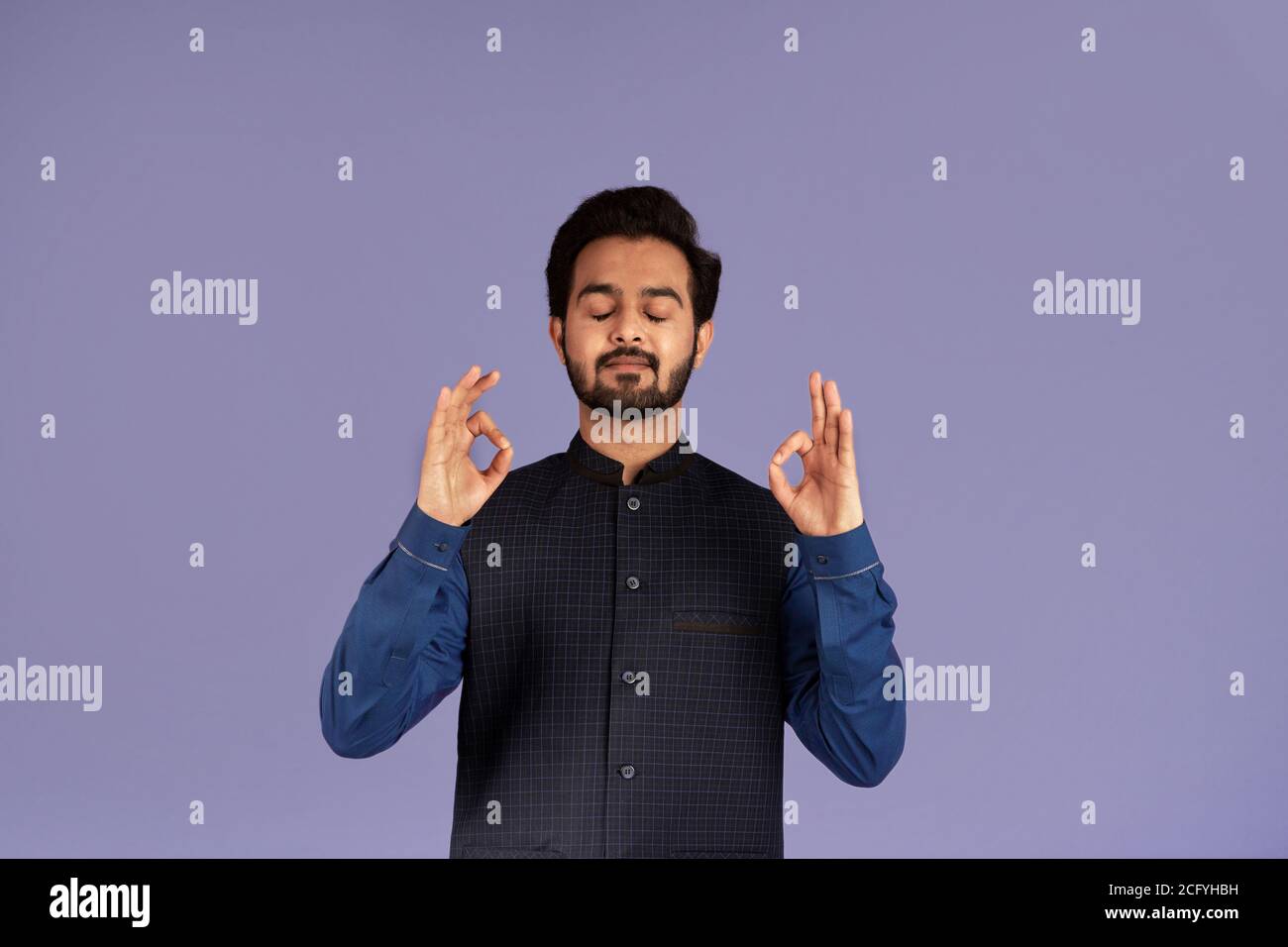 Friedlicher indischer Kerl meditiert und macht Yoga Mudras auf Flieder Hintergrund Stockfoto