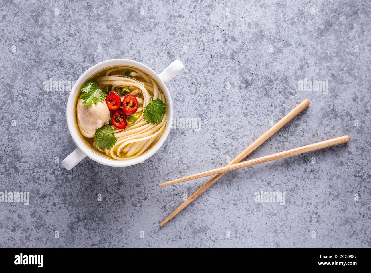 Würzige asiatische Suppe. Hühnerbouillon und Fleisch, Chili, Koriander, Frühlingszwiebeln und japanische Nudeln in weißer Schüssel mit hölzernen Essstäbchen auf grauer Brandung Stockfoto
