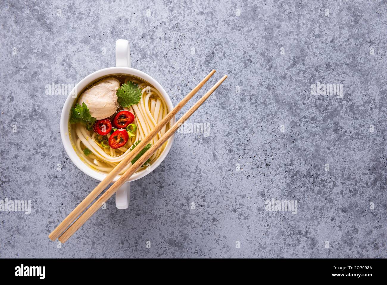 Würzige asiatische Suppe. Hühnerbouillon und Fleisch, Chili, Koriander, Frühlingszwiebeln, japanische Nudeln in weißer Schüssel auf grauer Oberfläche. Holzstäbchen auf Stockfoto