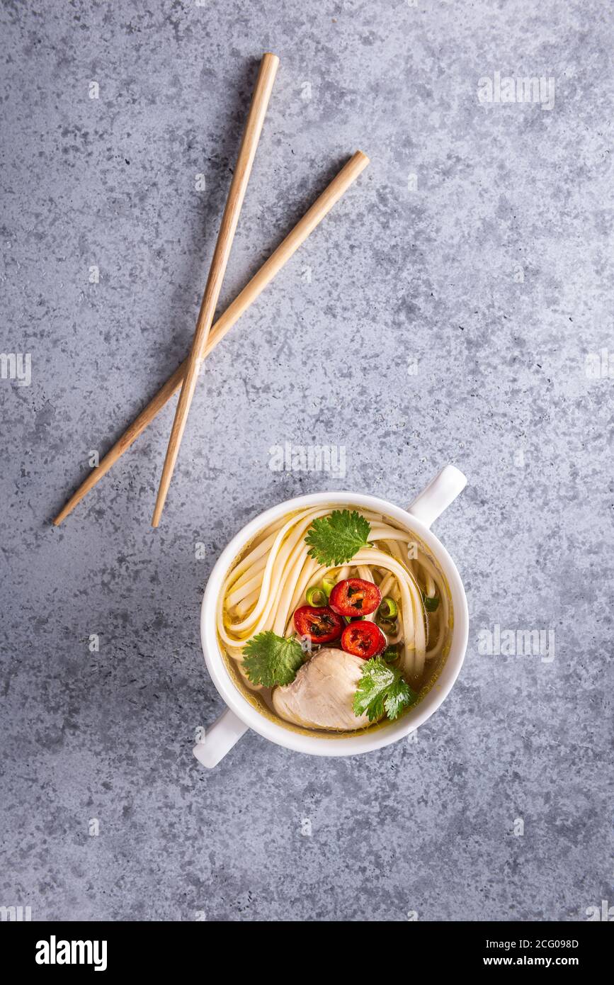 Würzige asiatische Suppe. Hühnerbouillon und Fleisch, Chili, Koriander, Frühlingszwiebeln und japanische Nudeln in weißer Schüssel mit hölzernen Essstäbchen auf grauer Brandung Stockfoto