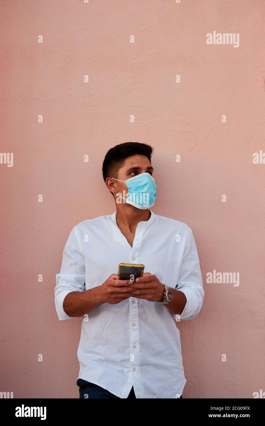 Junger latein mit Maske schaut auf sein Telefon Rosa Hintergrund Stockfoto