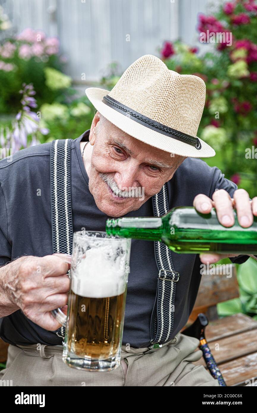 Älterer Mann in einem Hut trinkt Bier vor einer Kneipe auf einer Bank im  Garten. Der alte Mann gießt Bier in ein Glas Stockfotografie - Alamy