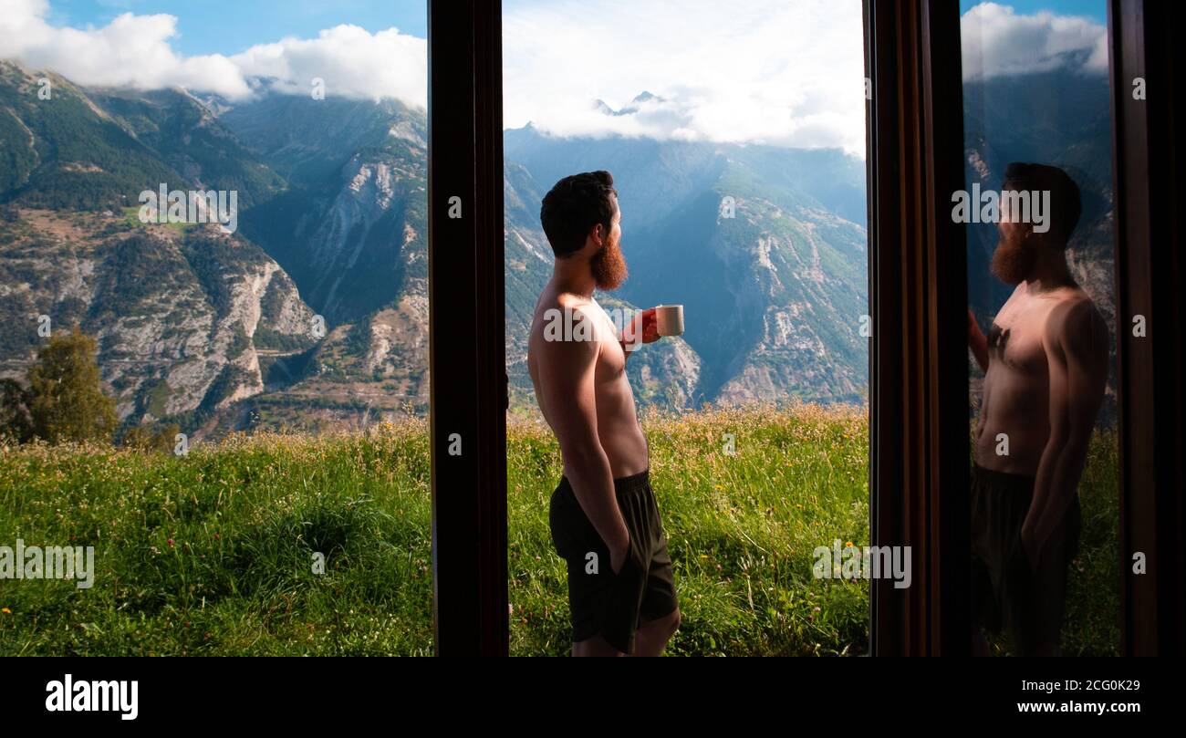 Junger Mann, der neben einem Fenster steht und seinen Morgenkaffee trinkt Mit schöner Berglandschaft in Österreich Stockfoto