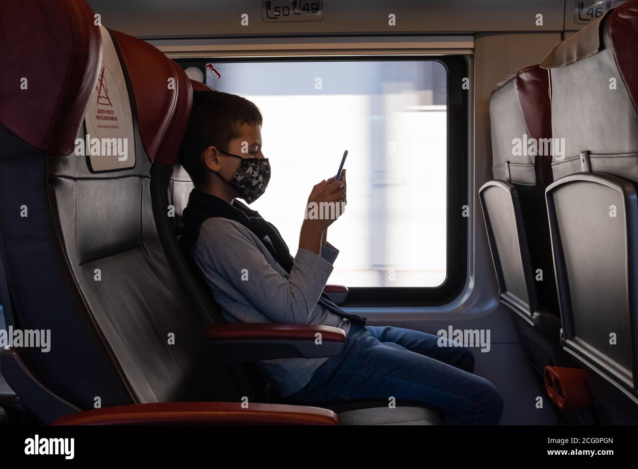 Kleiner Junge, der mit dem Zug unterwegs ist und ein Smartphone sucht. Kind sitzt am Fenster des Waggons und trägt eine schützende Gesichtsmaske. Stockfoto