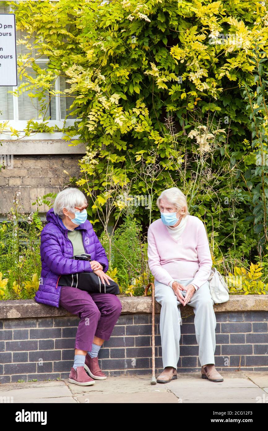 Zwei Frauen ältere Rentner tragen Gesichtsmasken sozial Distanziertes Sitzen an einer Wand, das während des Corona-Virus spricht Covid 19 Pandemie Stockfoto