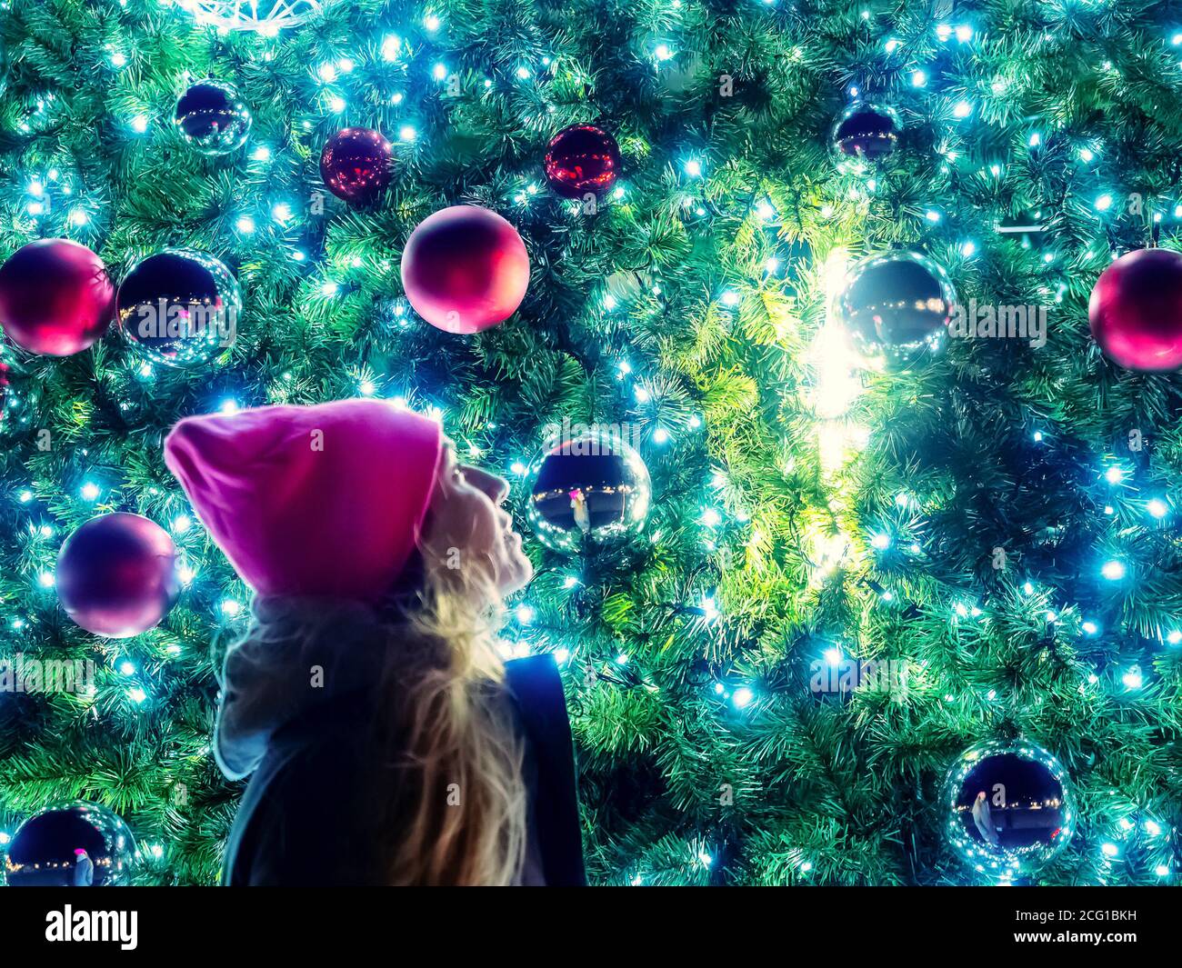Die Neujahrsbälle auf den Zweigen des Weihnachtsbaums mit einer Girlande und einem Mädchen, das daneben steht, spiegeln sich im Ball wider. Stockfoto